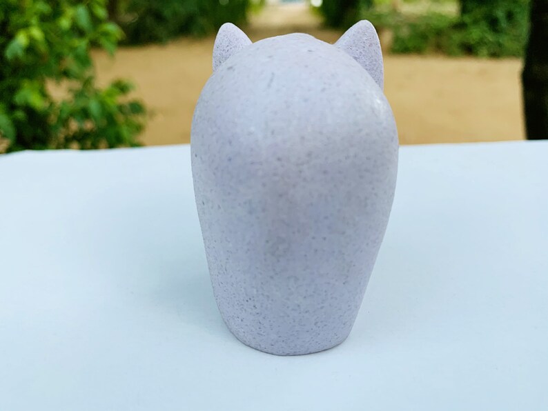 Może przedstawiać: Mała, jasnolilawa figurka ceramiczna w kształcie kota z zaokrąglonymi uszami. Figurka stoi na białym tle.