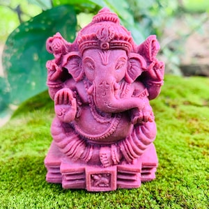 May include: A small, pink, detailed statue of the Hindu deity Ganesha. The statue is sitting on a base with intricate carvings.