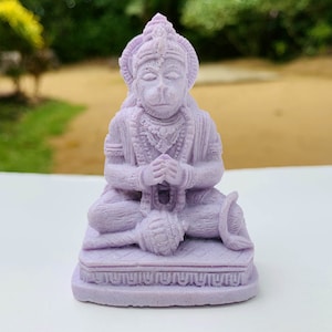 Puede incluir: Una pequeña estatua de un dios mono sentado de color púrpura, detallada, con las manos en posición de oración. La estatua está sobre una base con diseños intrincados.