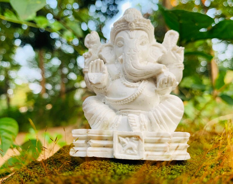 May include: White Ganesha statue with a trunk curled to the left, sitting on a tiered base. The statue is placed on a bed of moss and grass.