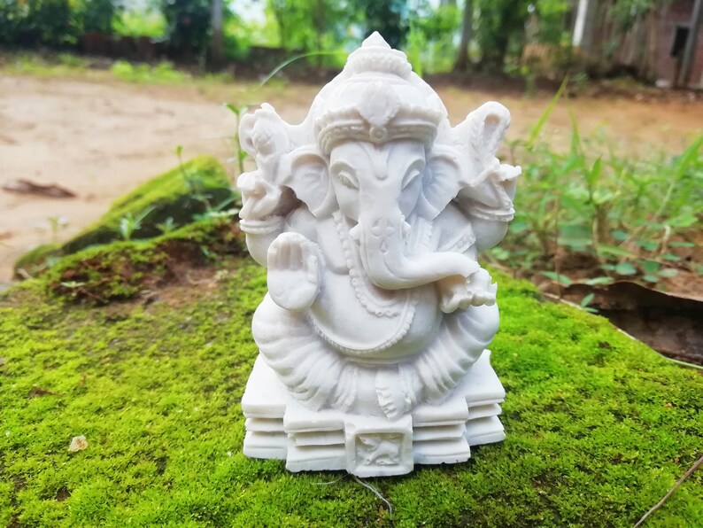 May include: White Ganesha statue with a detailed design, sitting on a tiered base. The statue is placed on a bed of green moss.