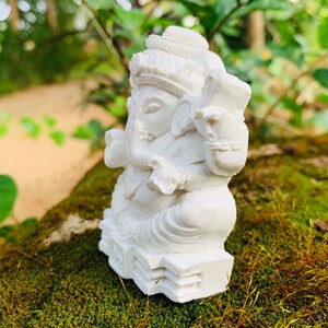 May include: White figurine of the Hindu deity Ganesha, sitting on a base with intricate details. The figurine is made of a smooth, white material and is placed on a bed of green moss.