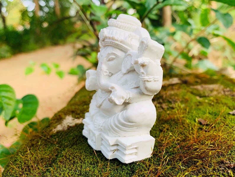 May include: White figurine of the Hindu deity Ganesha, sitting on a base with intricate details. The figurine is made of a smooth, white material and is placed on a bed of green moss.