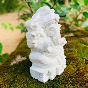 May include: White Ganesha statue with intricate details, sitting on a bed of moss. The statue is made of a smooth, white material and has a traditional design.