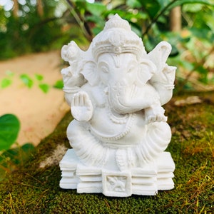 May include: White Ganesha statue with a detailed design, sitting on a stack of books. The statue is made of a smooth, white material and has a serene expression.