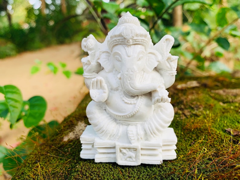 May include: White Ganesha statue with a detailed design, sitting on a stack of books. The statue is made of a smooth, white material and has a serene expression.