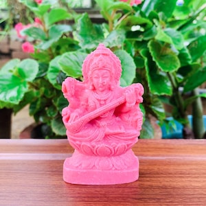 Puede incluir: Una figurita rosa de la diosa hindú Saraswati tocando un veena, un instrumento de cuerda. Está sentada sobre una flor de loto y tiene cuatro brazos. La figurita está hecha de un material liso y pulido.