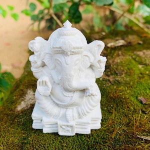 May include: White Ganesha statue with a detailed design, sitting on a base with multiple layers. The statue is placed on a bed of green moss.