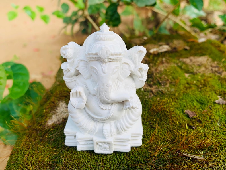 May include: White Ganesha statue with a detailed design, sitting on a base with multiple layers. The statue is placed on a bed of green moss.