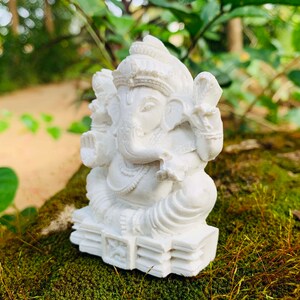 May include: White Ganesha statue with intricate details, sitting on a base with a decorative design. The statue is placed on a bed of green moss.