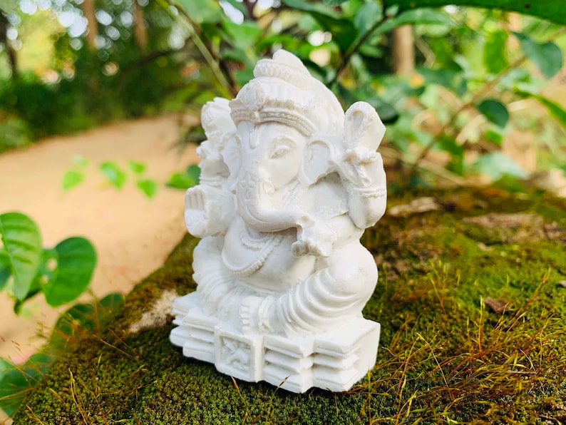 May include: White Ganesha statue with intricate details, sitting on a base with a decorative design. The statue is placed on a bed of green moss.
