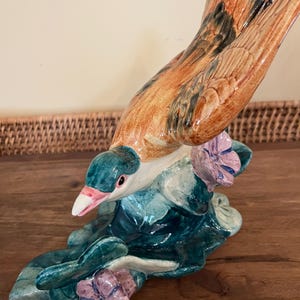 May include: A ceramic bird figurine with brown, green, and blue painted details. The bird is perched on a green leafy base with purple flowers.