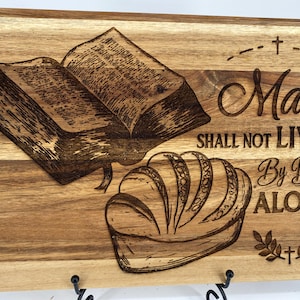 Könnte beinhalten: Holzschneidebrett mit einem eingravierten Design, das ein offenes Buch und ein Brotlaib zeigt. Der Text "Man Shall Not Live By Bread Alone" ist ebenfalls auf dem Brett eingraviert. Das Brett hat eine natürliche Holzmaserung.