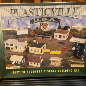May include: A Plasticville U.S.A. O scale building kit box featuring a printed illustration of a miniature town layout with various houses, a gas station, and small toy vehicles. The box includes text labels such as Realistic Detailing, Parts Molded in Color, Snap Fit Assembly, and No Glue Necessary.