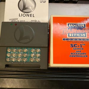 May include: A gray Lionel Trainmaster SC-1 switch and accessory controller with a terminal block featuring multiple screw terminals. The device is accompanied by its original orange and white box and a manual with the Lionel logo printed on it.
