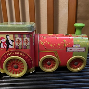 May include: A metal tin shaped like a train engine, colored in red and light green. The side features illustrations of penguins and the text Santa Express. It has a green lid, a green chimney, and four wheels with red spokes and green rims.