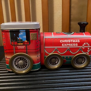 May include: A red and green metal train-shaped tin container with a silver lid. The side features the text "CHRISTMAS EXPRESS" in white lettering, a candy cane graphic, and a window showing a silhouette of a person and wrapped gifts.