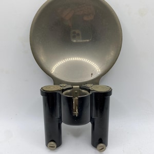 May include: A vintage automotive part featuring a large, circular, silver-toned metal component connected to a black, cylindrical base. The base has two vertical cylinders with screw-like details.