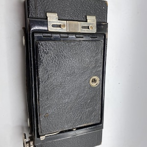 May include: A vintage black camera with a textured leather exterior. The camera features a silver metal clasp and a lens cover. The camera is rectangular and has a small handle on the side. The camera is likely from the early to mid-20th century.