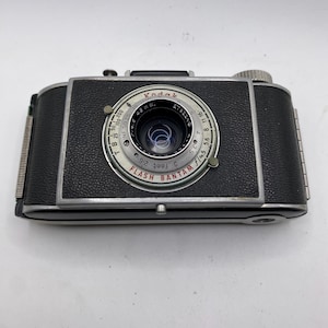 Puede incluir: Una cámara Kodak Flash Bantam vintage. La cámara es negra con una lente y un dial plateados. La lente tiene el texto "Kodak" y "Flash Bantam". La cámara tiene una perilla de cuerda plateada y un botón plateado.