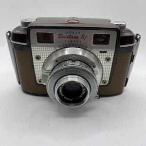 Puede incluir: Cámara Kodak Bantam RF vintage. La cámara tiene un cuerpo marrón y plateado con una lente negra. La lente dice "Kodak Ektanon Lens 50 mm f/3.9". La cámara tiene las palabras "Kodak Bantam RF Camera" impresas en la parte delantera.