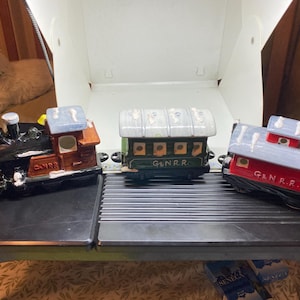 May include: Three ceramic train cars: a black and brown locomotive, a green passenger car, and a red caboose. Each car has a light blue roof and the letters "G & N.R.R." or "CANRR" painted on them. The train cars are on a black surface.