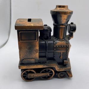 Op de afbeelding: Een gedetailleerde, antieke puntenslijper in de vorm van een trein. De puntenslijper is gemaakt van metaal met een bronzen en zwarte afwerking. De trein heeft wielen, een schoorsteen en een klein compartiment bovenop.