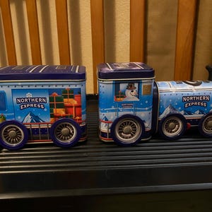 May include: Two blue metal train-shaped tins featuring the text Northern Express. The locomotive and carriage are decorated with snowy mountain scenes, gift boxes, and a snowman. Each piece has silver-toned wheels and a dark blue lid.