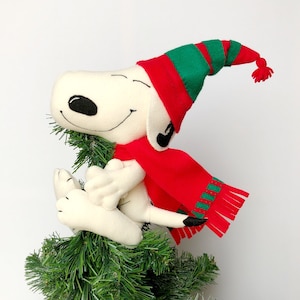 Könnte beinhalten: Ein weißer Plüsch-Snoopy, der einen rot-grünen Weihnachtsmann-Hut und einen roten Schal mit grünem Besatz trägt, sitzt auf einem grünen Weihnachtsbaum.