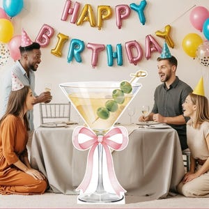 May include: A large, die-cut cardboard cutout of a martini glass filled with a pale yellow liquid and a skewer of three green olives. The stem of the glass is decorated with a large, light pink ribbon bow tied in a decorative knot.