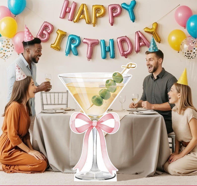 May include: A large, die-cut cardboard cutout of a martini glass filled with a pale yellow liquid and a skewer of three green olives. The stem of the glass is decorated with a large, light pink ribbon bow tied in a decorative knot.