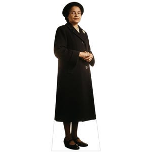 May include: A life-size cardboard cutout of Rosa Parks standing and facing forward. She is wearing a long black coat, a matching black hat, and dark-rimmed glasses. Her hands are clasped in front of her, and she wears black shoes on her feet.