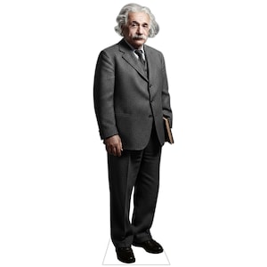 May include: A life-sized cardboard cutout of Albert Einstein standing in a grey suit, white shirt, and dark tie. The figure has white hair, a mustache, and holds a brown book under one arm. The cutout is mounted on a white base for stability.