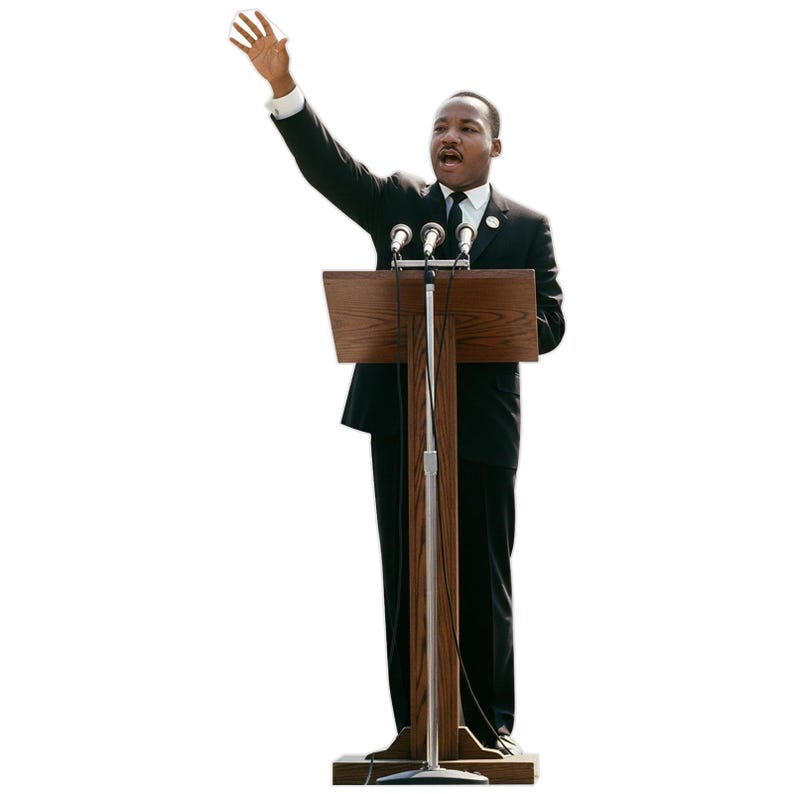 May include: A cutout of Martin Luther King Jr. wearing a black suit, white shirt, and black tie, standing behind a wooden podium with three microphones. One arm is raised with an open palm, and the mouth is open as if speaking to an audience.