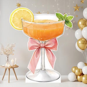 May include: A large cutout of a cocktail glass filled with an orange liquid, garnished with a bright yellow lemon slice and green mint leaves. The stem of the glass is decorated with a large, soft pink ribbon tied into a bow at the center.
