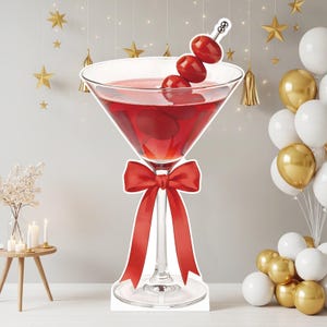May include: A large martini glass cutout featuring a red liquid, three red olives on a silver cocktail pick, and a bright red ribbon tied around the stem. The glass is clear with a wide bowl and a slender stem, standing on a circular base.