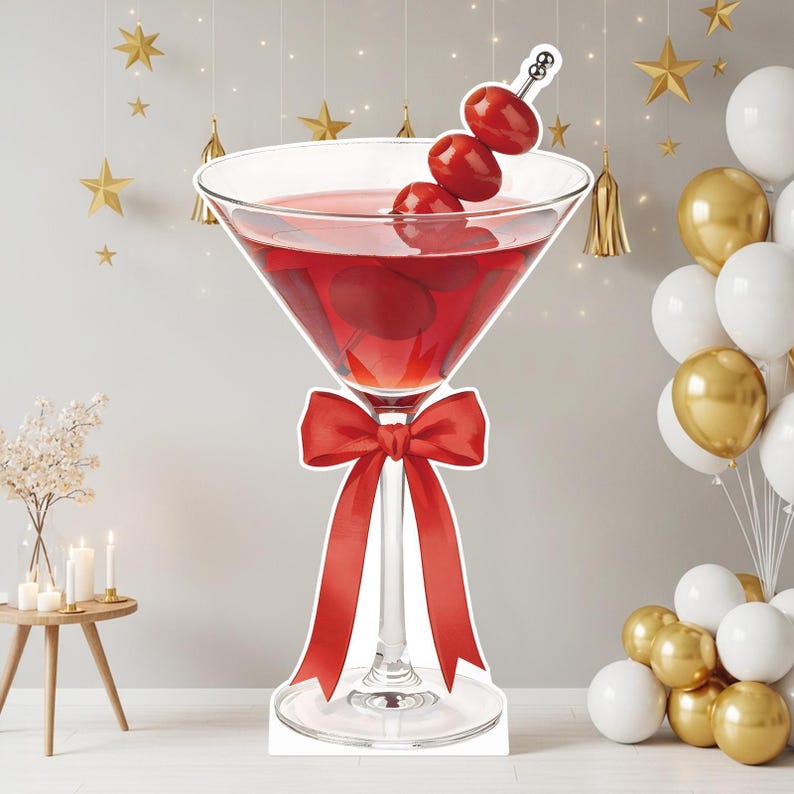 May include: A large martini glass cutout featuring a red liquid, three red olives on a silver cocktail pick, and a bright red ribbon tied around the stem. The glass is clear with a wide bowl and a slender stem, standing on a circular base.