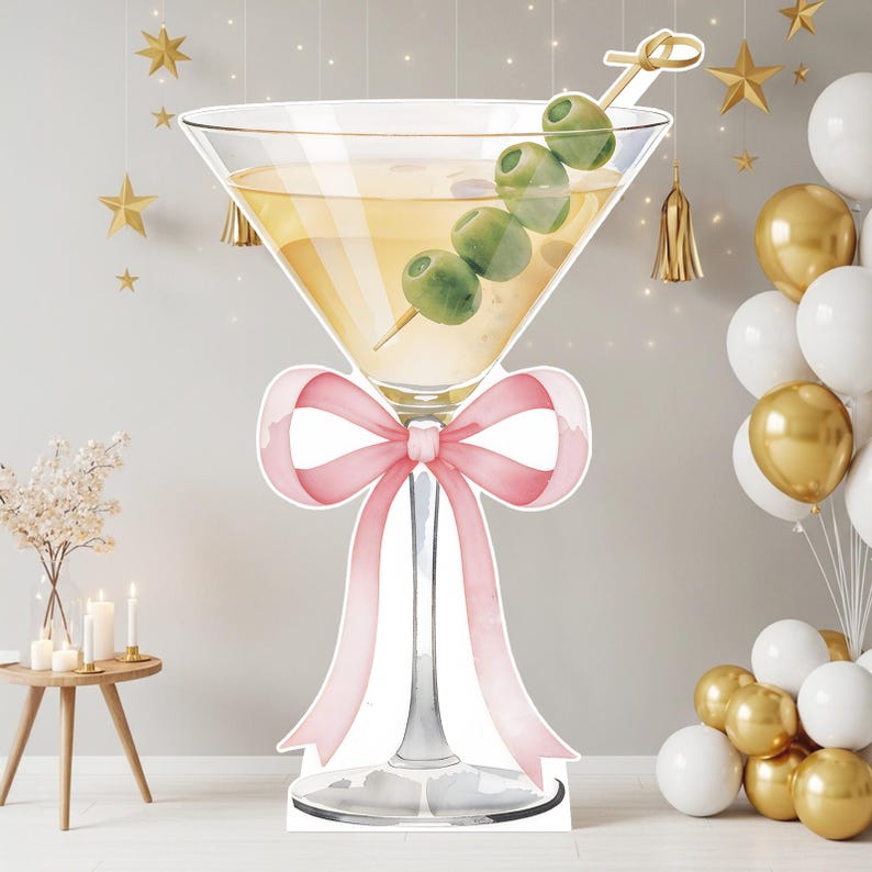 May include: A large cutout of a martini glass filled with a pale yellow liquid and three green olives on a cocktail pick. The stem of the glass is decorated with a soft pink ribbon tied in a bow. The design features a white outline border.