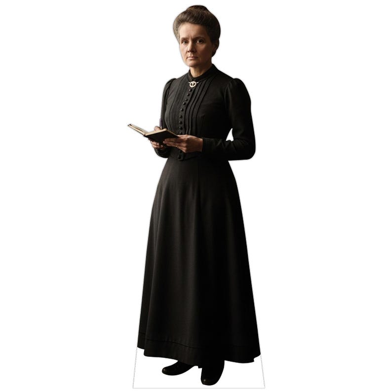 May include: A life-sized cardboard cutout of Marie Curie standing in a long-sleeved black dress with a high collar, vertical pleats, and a row of buttons down the front. The figure holds a small open notebook and pen, with hair styled in a bun.