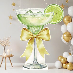 May include: A large cutout of a margarita glass filled with a light green liquid and ice cubes, featuring a salt-rimmed edge and a lime slice garnish. A prominent yellow ribbon bow is tied around the stem of the glass, creating a decorative display.