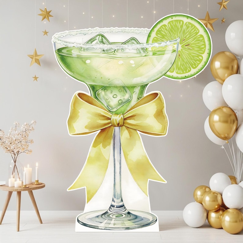 May include: A large cutout of a margarita glass filled with a light green liquid and ice cubes, featuring a salt-rimmed edge and a lime slice garnish. A prominent yellow ribbon bow is tied around the stem of the glass, creating a decorative display.