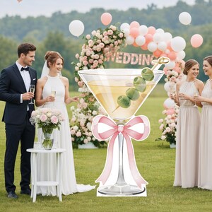 May include: A large, flat, die-cut cardboard cutout of a martini glass filled with a yellow liquid and three green olives on a cocktail pick. The glass stem is decorated with a large, light pink ribbon bow at the base of the bowl.