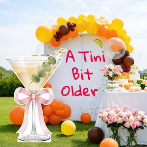 May include: A large cutout of a martini glass with a pink bow and an olive garnish, positioned in front of a white backdrop featuring the text A Tini Bit Older in pink, surrounded by orange, yellow, and brown balloons and floral arrangements.
