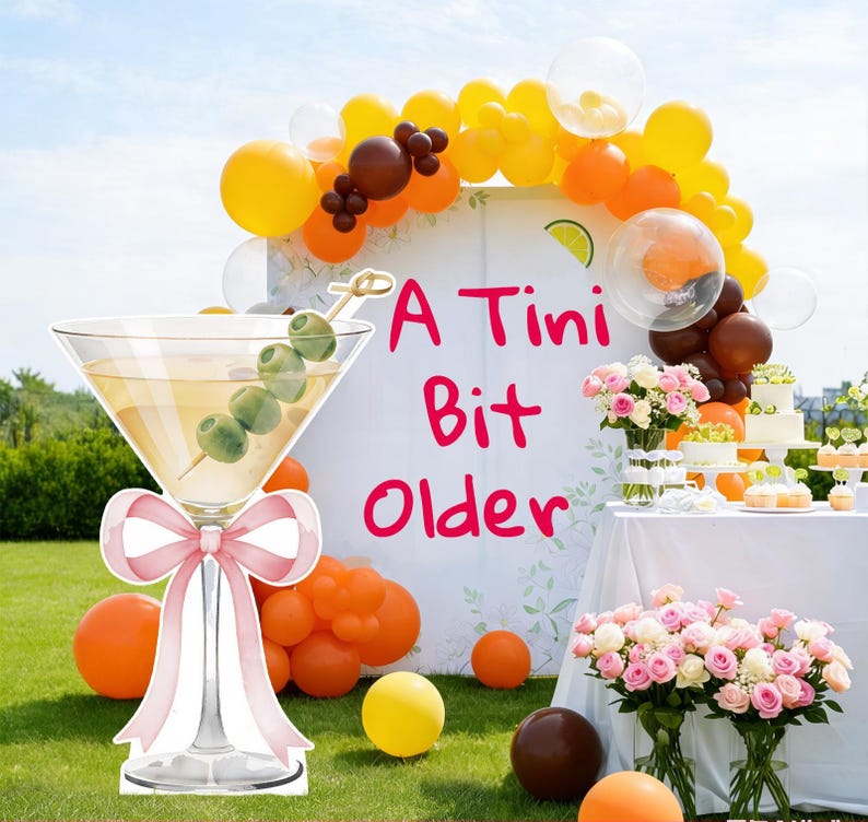 May include: A large cutout of a martini glass with a pink bow and an olive garnish, positioned in front of a white backdrop featuring the text A Tini Bit Older in pink, surrounded by orange, yellow, and brown balloons and floral arrangements.