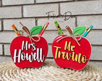 May include: Two red apple-shaped desk organizers with green leaves and pencils. One has "Mrs. Howell" in white script, the other "Mr. Trevino" in yellow. A perfect teacher gift.