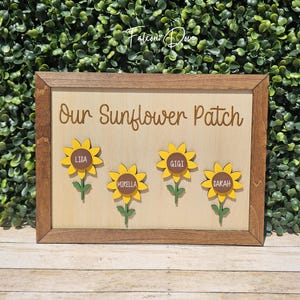 May include: A rectangular wooden sign with a dark brown frame and a light wood background. The text "Our Sunflower Patch" is printed in brown. Four yellow sunflowers with green stems are attached, each featuring a brown center with the names Lisa, Mirella, Gigi, and Sarah.