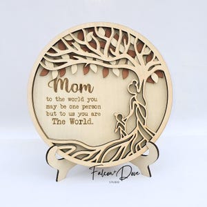 May include: A wooden, layered circular plaque with a tree design and the word "Mom" in script. The plaque includes the text "to the world you may be one person but to us you are The World." It is displayed on a wooden stand.