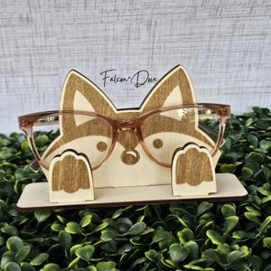 May include: A light-colored wooden eyeglass stand shaped like a fox head with two raised paws holding a pair of brown-rimmed glasses. The stand features the text Falcon + Dove Studio printed in black ink above the fox's ears on the wooden surface.