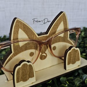 May include: A light-colored wooden eyeglass holder shaped like a fox head with two small paw-shaped supports at the base. The holder features dark brown engraved details and the text Falcon + Dove Studio. A pair of light brown glasses rests on it.
