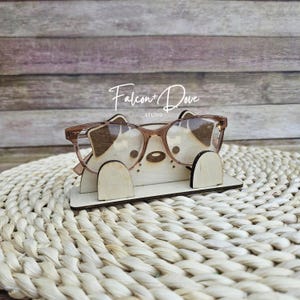 May include: A light-colored wooden eyeglass stand shaped like a cat face with two small ears and a nose, holding a pair of brown-rimmed glasses. The stand features the text Falcon + Dove STUDIO printed in white above the glasses.
